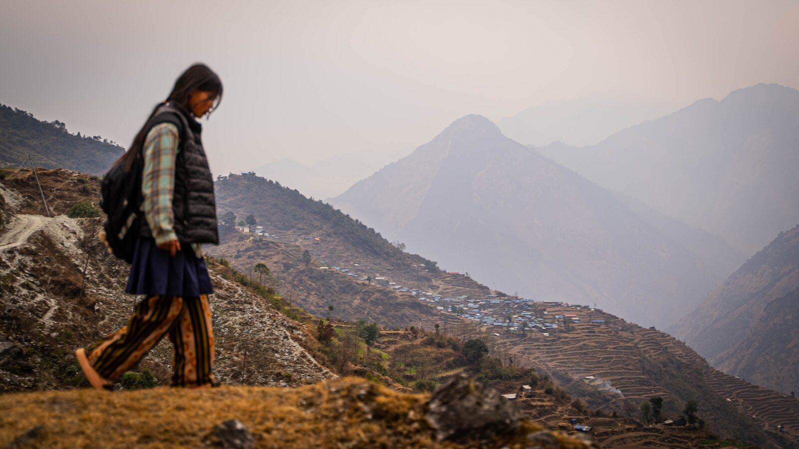 Una persona con mochila camina por un sendero de montaña en Nepal, con una aldea y cultivos en terrazas al fondo.